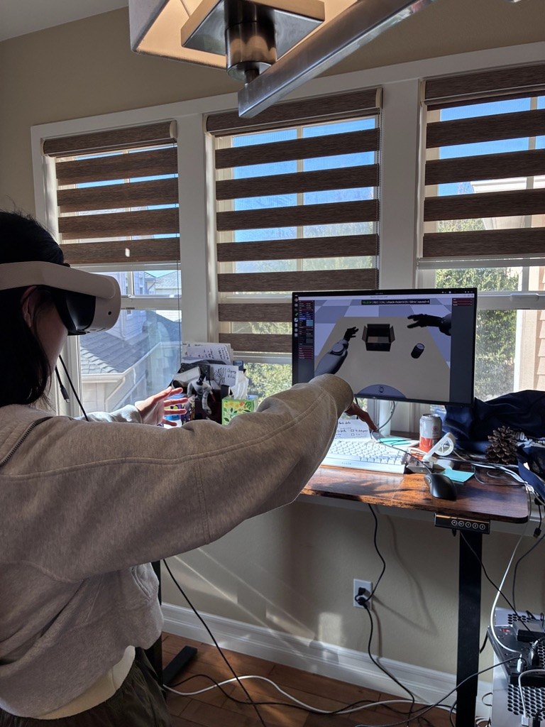 Wearing a VR headset with both arms raised, teleoperating a two-armed robot in simulation; the monitor shows the simulated hands and the objects being manipulated